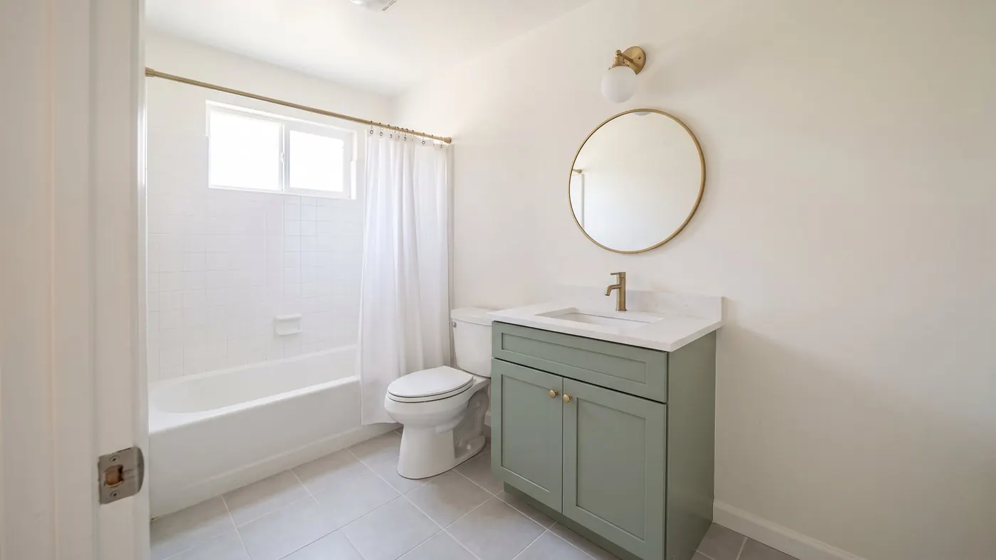 Design rendering of a hall bathroom with a new sage green vanity, a white toilet, a brushed brass faucet, round mirror and wall light, and the original tub with white tile and a white curtain