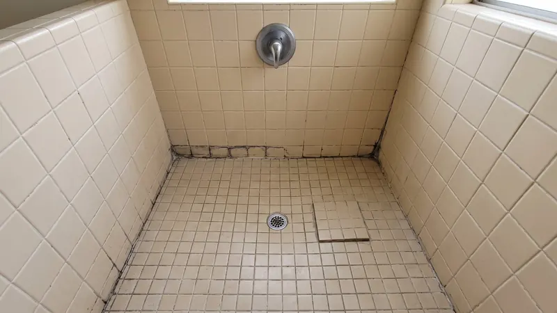 Design rendering of a dated tiled shower stall with cracked, darkened grout along the floor and a loose floor tile near the drain