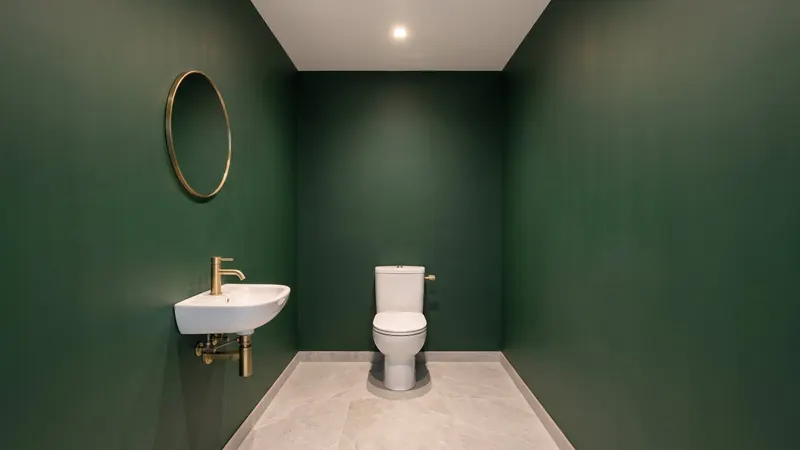 Design rendering of a powder room with deep green painted walls, a small white wall mounted sink with a brass faucet and exposed brass trap, a round brass mirror and a white toilet on the back wall