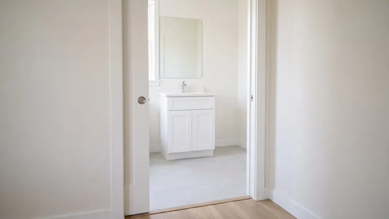 Design rendering of a small bathroom seen from a hallway through an open pocket door slid most of the way into the wall, with a white vanity and a plain mirror inside