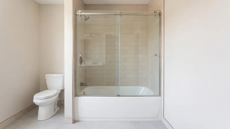 Design rendering of a small bathroom with a white tub behind two clear sliding glass panels hung from a brushed nickel top track, sand colored wall tile and a white toilet beside the alcove
