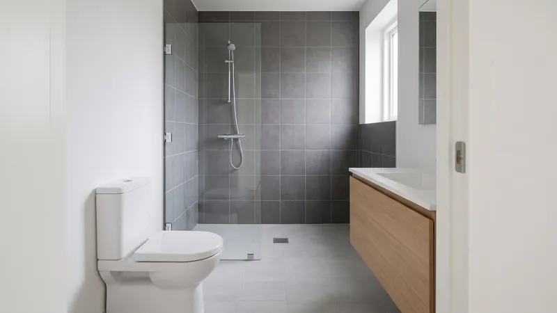 Design rendering of a small bathroom where a walk-in shower with charcoal wall tile and one clear glass panel fills the far end, with a white toilet on one side and a floating light oak vanity on the other