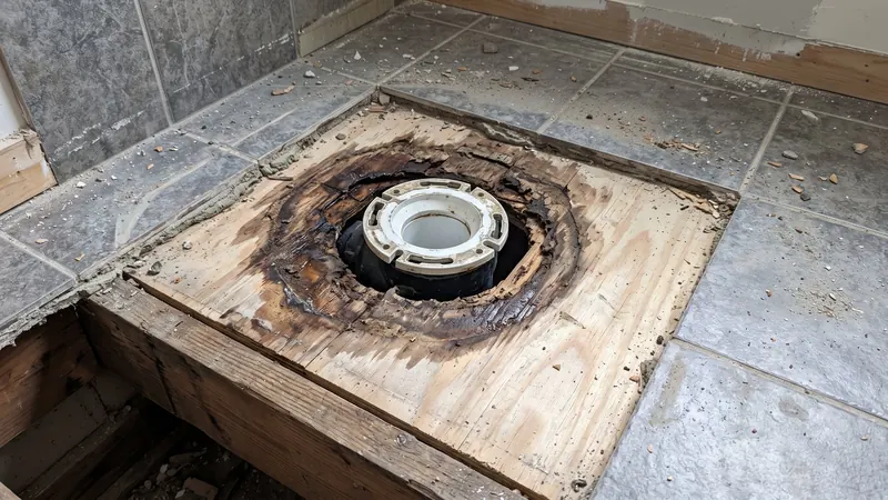 Design rendering of a bathroom floor opened around a toilet flange, showing dark water stained plywood subfloor and a floor joist at the edge